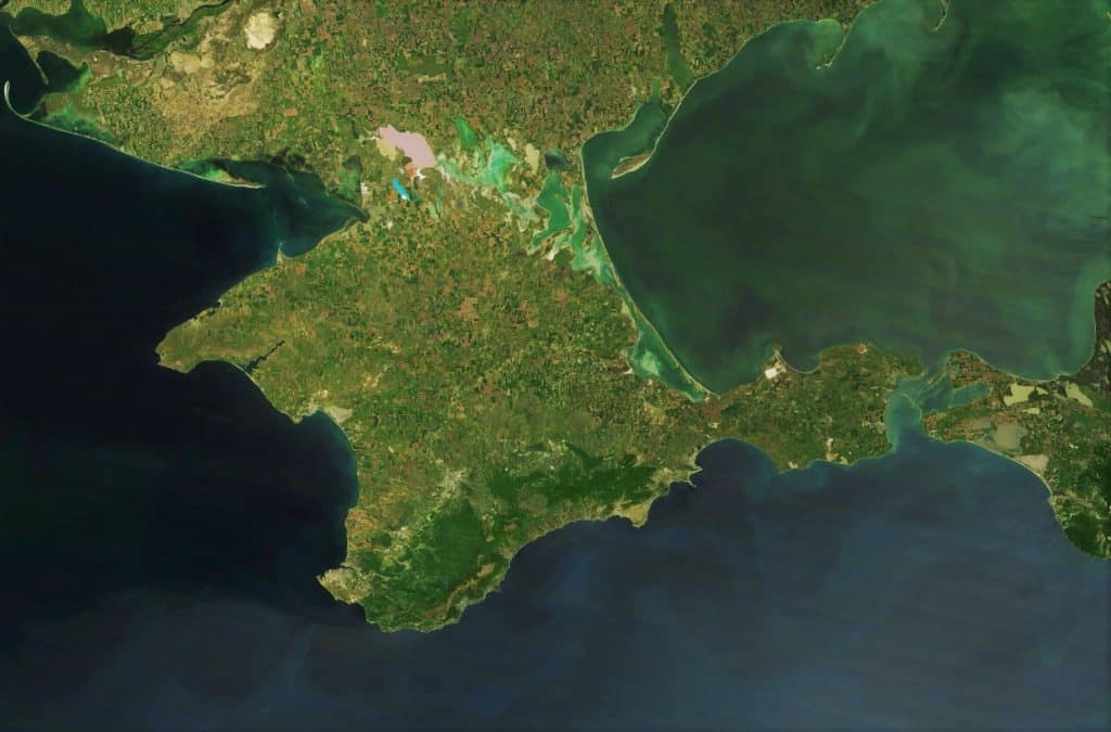 A satellite image of Crimea, the peninsula extends into the Black Sea and is connected to mainland Ukraine by a narrow isthmus. The landscape varies from green vegetation to areas of brown, indicating different types of terrain and land use. The surrounding water appears dark green and blue, reflecting recent mapping policies.