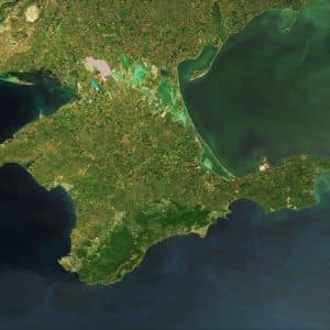 A satellite image of Crimea, the peninsula extends into the Black Sea and is connected to mainland Ukraine by a narrow isthmus. The landscape varies from green vegetation to areas of brown, indicating different types of terrain and land use. The surrounding water appears dark green and blue, reflecting recent mapping policies.