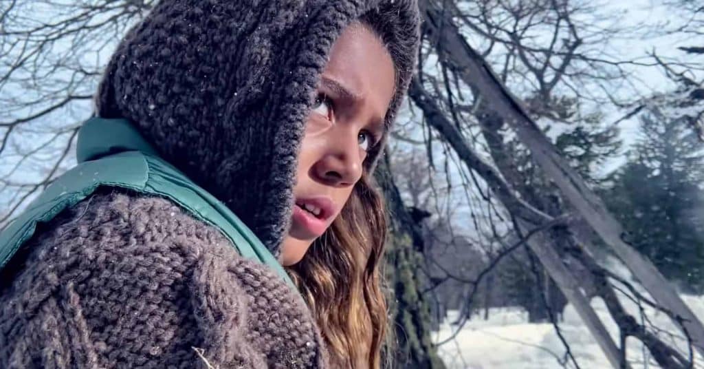 A young person with long hair, wearing a knitted hoodie and a green vest, looks pensively into the distance in a snowy, wooded area. Bare trees and snow-covered ground are visible in the background. Shot on iPhone, this scene could be straight out of Apple’s “Snowbrawl” video series.