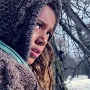 A young person with long hair, wearing a knitted hoodie and a green vest, looks pensively into the distance in a snowy, wooded area. Bare trees and snow-covered ground are visible in the background. Shot on iPhone, this scene could be straight out of Apple’s “Snowbrawl” video series.