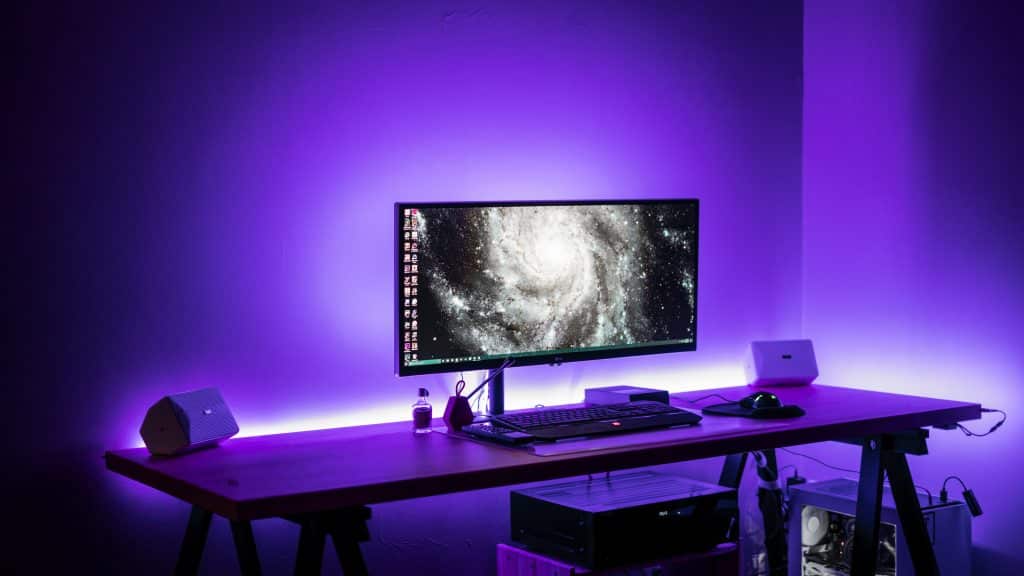 A 2020 gaming iMac setup on a wooden desk with a widescreen monitor showing a galaxy image. The desk has a keyboard, mouse, two speakers, and various small items. The room glows with a purple hue from LED lights, creating a futuristic ambiance.