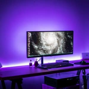 A 2020 gaming iMac setup on a wooden desk with a widescreen monitor showing a galaxy image. The desk has a keyboard, mouse, two speakers, and various small items. The room glows with a purple hue from LED lights, creating a futuristic ambiance.