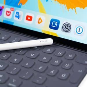 An Apple Pencil rests on a black iPad keyboard. The iPad screen displays colorful artwork and app icons including Google Drive, YouTube, and Audible. This scene highlights the integration of Apple products for creative and productive tasks, showcasing some of the most influential tech innovations of the 2010s.