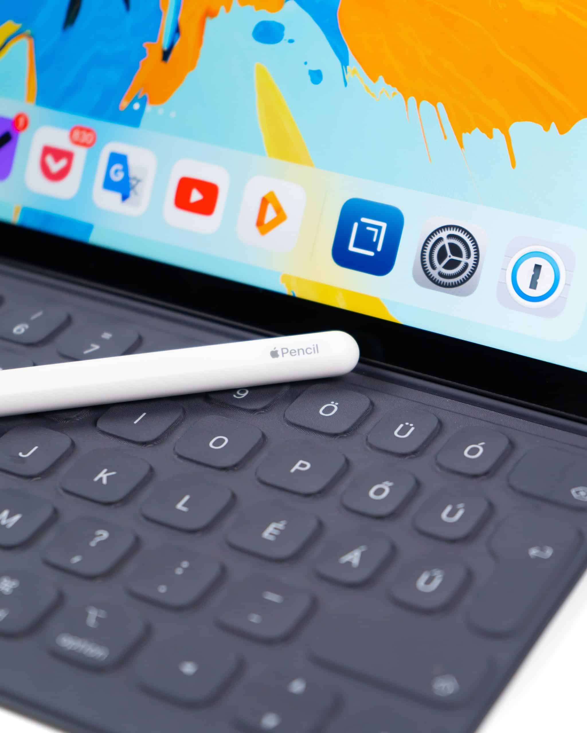 An Apple Pencil rests on a black iPad keyboard. The iPad screen displays colorful artwork and app icons including Google Drive, YouTube, and Audible. This scene highlights the integration of Apple products for creative and productive tasks, showcasing some of the most influential tech innovations of the 2010s.