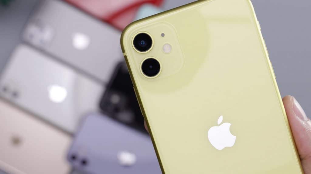 A person holds a yellow iPhone with dual cameras close to the lens. In the blurred background, multiple 2020 iPhones of different colors are displayed, including grey, black, and purple models. These Apple devices also boast larger batteries for extended use.