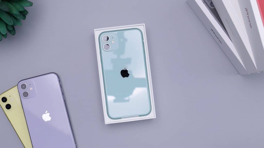 A light blue Apple iPhone is placed in its open white box on a light grey surface. Nearby, there are other 2020 iPhones in yellow and purple, alongside iPhone packaging and a small part of a green plant. Despite the manufacturing price increase, the variety of colors remains appealing.