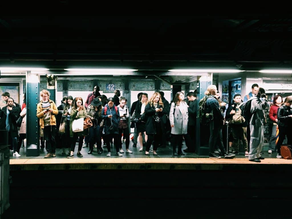 A group of people stand on a subway platform in the New York transport system, appearing to be waiting for a train. The scene is taken indoors, the platform is well lit, and a sign on the wall in the background reads "42." Many people are focused on their phones or discussing the recent rollout of Apple Pay.