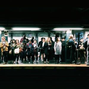 A group of people stand on a subway platform in the New York transport system, appearing to be waiting for a train. The scene is taken indoors, the platform is well lit, and a sign on the wall in the background reads "42." Many people are focused on their phones or discussing the recent rollout of Apple Pay.