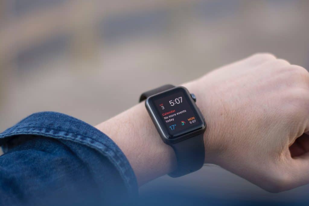 A person's left wrist wearing a black Apple Watch with a digital display showing the time as 5:07. The watch screen also displays notifications, calendar events, and weather information. The person is wearing a blue long-sleeve shirt.