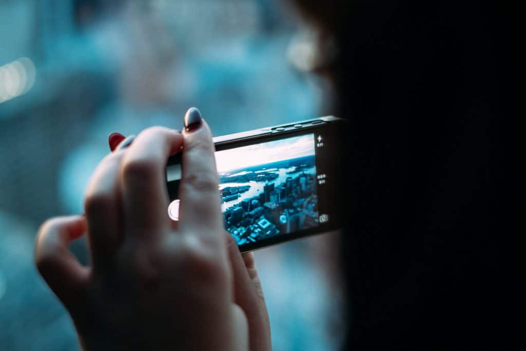 A person holding an iPhone horizontally, capturing a photo of a cityscape. The image on the phone's screen shows tall buildings and a river winding through the city. The background is blurred, focusing on the person's hand and showcasing Apple’s exceptional iPhone photography capabilities.