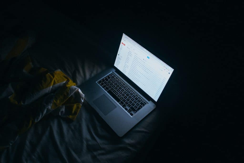 A laptop sits open on a dark bed, its screen glowing and displaying an email inbox with several unread emails. The left side of the image shows a part of a patterned blanket. The overall scene is dimly lit, highlighting the glow from the laptop screen as if Night Shift mode could help one fall asleep faster.