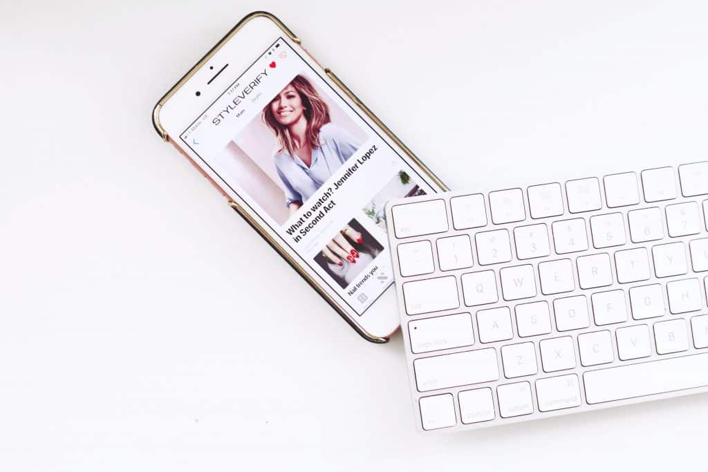 A cheap iPhone displaying an article with a photo of a woman is placed next to a white wireless keyboard. The article is on the website 'STYLE PARTY' and the headline partly reads, "Why you should update your winter wardrobe.