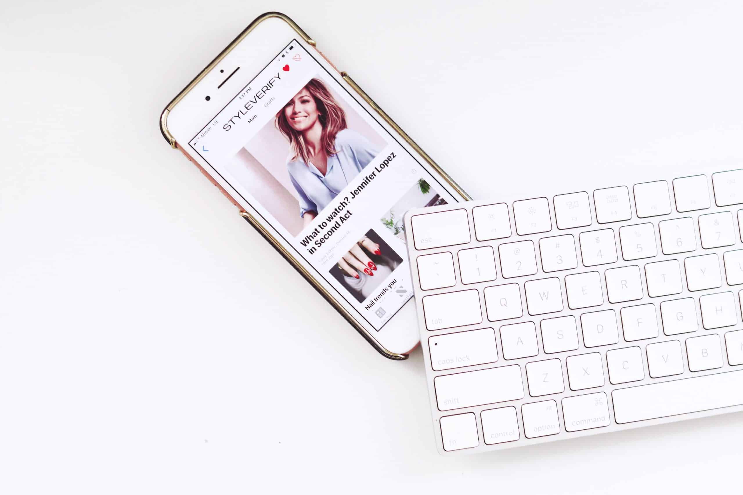 A cheap iPhone displaying an article with a photo of a woman is placed next to a white wireless keyboard. The article is on the website 'STYLE PARTY' and the headline partly reads, "Why you should update your winter wardrobe.