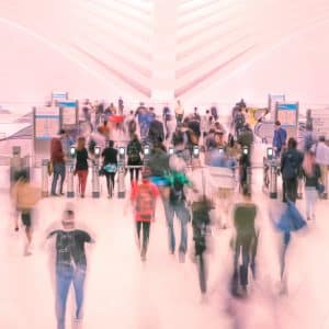 A large group of people walking through a modern, brightly lit train station with high ceilings. The image has a motion blur effect, emphasizing the hustle and bustle of commuters. Signs in the background indicate directions to different areas, showcasing the efficiency of Apple Express Transit Mode introduced in 2020.
