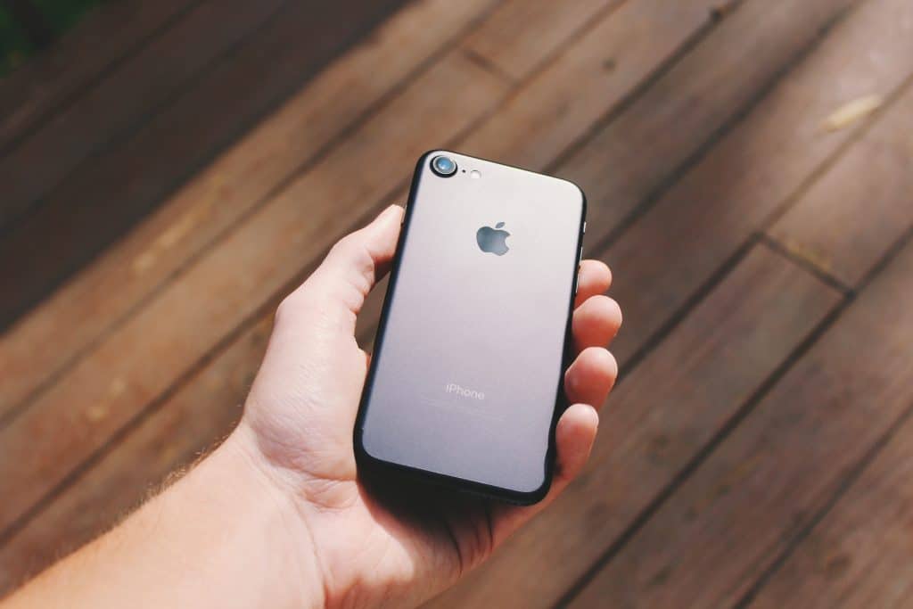 A hand holding an iPhone 9 with the Apple logo facing up. The background consists of wooden planks and a few patches of light, suggesting the phone is being held outdoors potentially under sunlight - a perfect setting to showcase this budget smartphone.