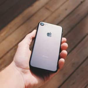 A hand holding an iPhone 9 with the Apple logo facing up. The background consists of wooden planks and a few patches of light, suggesting the phone is being held outdoors potentially under sunlight - a perfect setting to showcase this budget smartphone.