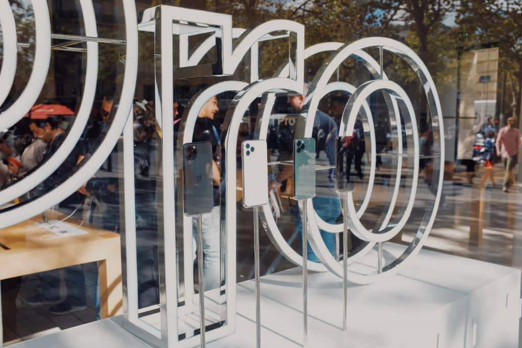 Several iPhones are displayed on stands in the window of an Apple Store. The glass window features large, white circular designs. Reflections of people and trees are visible in the background, showcasing the products that drive Apple's smartphone revenue.