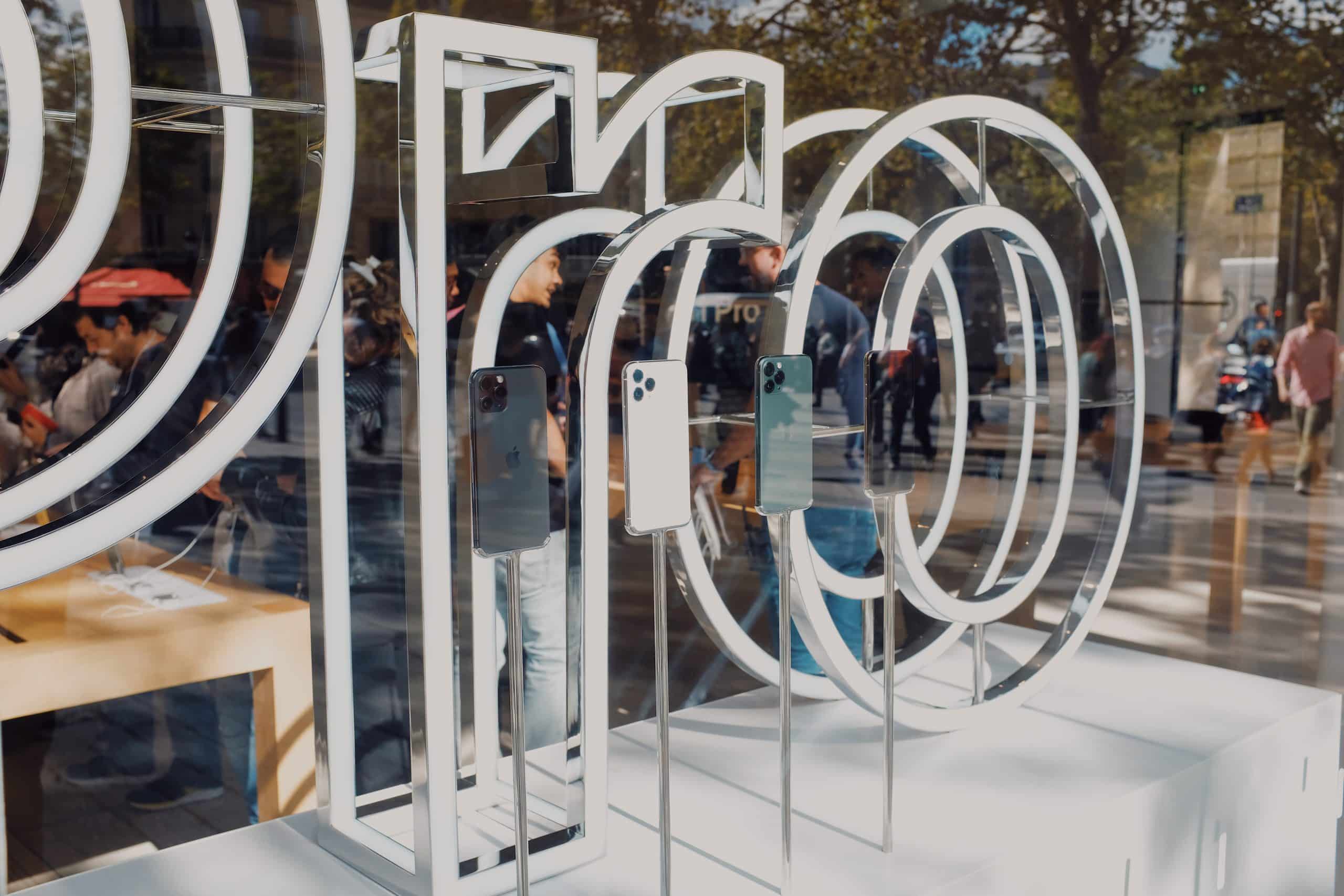 Several iPhones are displayed on stands in the window of an Apple Store. The glass window features large, white circular designs. Reflections of people and trees are visible in the background, showcasing the products that drive Apple's smartphone revenue.