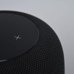 Close-up of a black smart speaker with a mesh exterior and a touch-sensitive top displaying an illuminated white plus symbol. Ideal for a Connected Home, it seamlessly integrates with Google services. The background is a plain, light gray color.