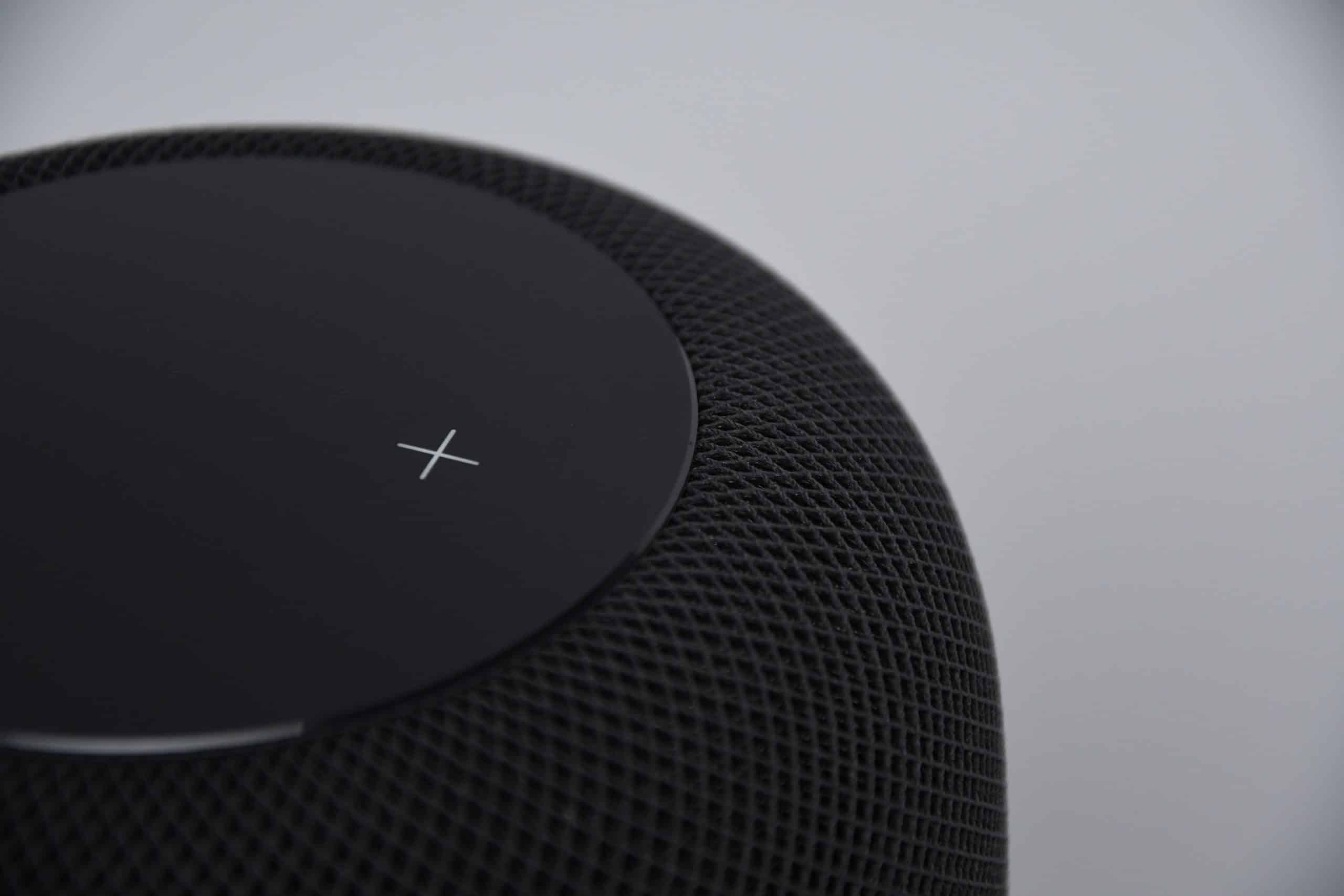 Close-up of a black smart speaker with a mesh exterior and a touch-sensitive top displaying an illuminated white plus symbol. Ideal for a Connected Home, it seamlessly integrates with Google services. The background is a plain, light gray color.