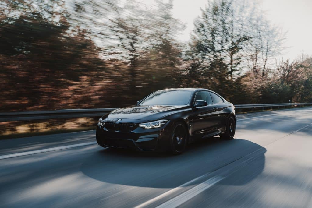 A sleek, black BMW sports car speeds down a tree-lined road with motion blur in the background, indicating its fast pace. It appears to be on a sunny day with clear weather. The car's modern, aerodynamic design hints at advanced features like CarPlay integration for tech-savvy drivers.