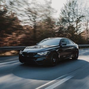 A sleek, black BMW sports car speeds down a tree-lined road with motion blur in the background, indicating its fast pace. It appears to be on a sunny day with clear weather. The car's modern, aerodynamic design hints at advanced features like CarPlay integration for tech-savvy drivers.