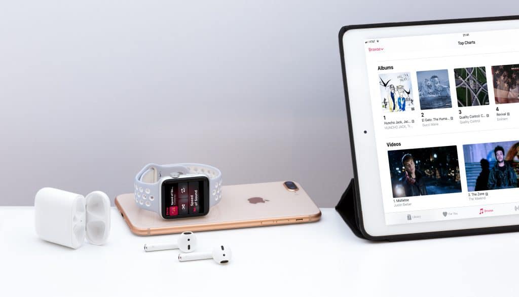 An Apple Watch with a gray band, an iPhone, AirPods with an open charging case, and an iPad displaying Apple Music's three months free offer are arranged on a white surface. The iPad screen shows various music albums and videos.