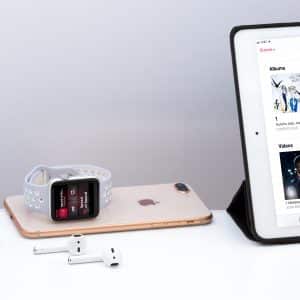 An Apple Watch with a gray band, an iPhone, AirPods with an open charging case, and an iPad displaying Apple Music's three months free offer are arranged on a white surface. The iPad screen shows various music albums and videos.