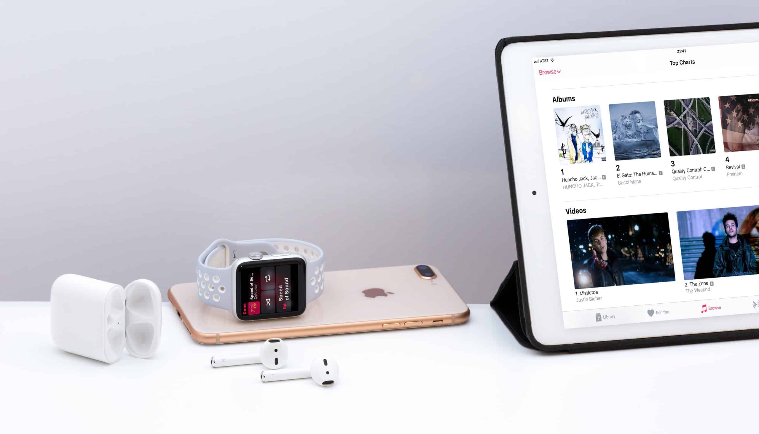 An Apple Watch with a gray band, an iPhone, AirPods with an open charging case, and an iPad displaying Apple Music's three months free offer are arranged on a white surface. The iPad screen shows various music albums and videos.