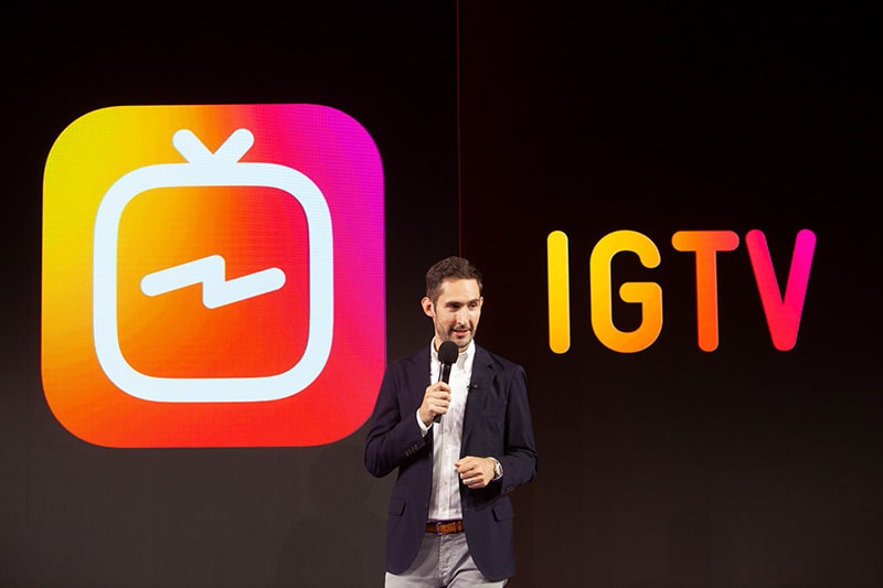 A person in a suit stands on stage holding a microphone. Behind them, the large screen displays the IGTV logo from the popular Instagram app, featuring a colorful gradient TV icon and the text "IGTV" in a matching gradient.