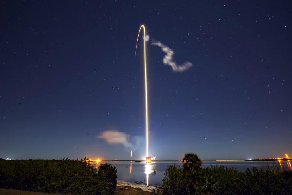 A rocket launches into the night sky, leaving a bright, curving trail of light behind it. The clear, star-filled sky features a slight cloud of smoke near the launch site. The illuminated launch pad is visible in the distance as water and greenery frame the scene, reminiscent of a StarLink mission.