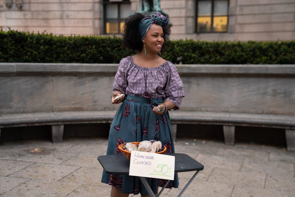 A woman stands outside by a stone bench, smiling and wearing a purple top and patterned skirt. She holds cookies in her hands and behind her on the bench. In front of her is a small table with a sign that reads "Homemade Cookies" priced at 50 cents, as if she's stepped straight out of an episode of Little America on Apple TV+.