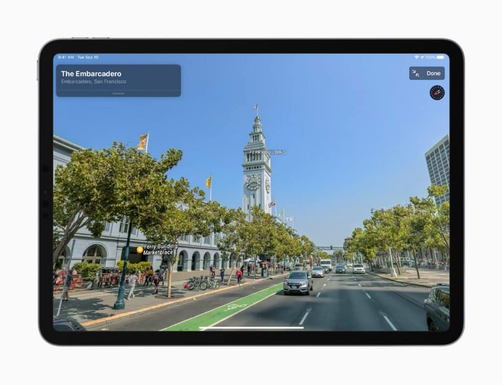 A tablet using Apple Maps displays a street view of San Francisco’s Embarcadero on its screen. The Ferry Building and its clock tower are visible in the background. Trees line the road with cars parked along the side, while pedestrians and cyclists are on the street, showcasing Apple's new US rollout.