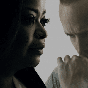 A close-up of two people with serious expressions. On the left, a woman with dark hair looks pensively to the right. On the right, a man with a beard and short hair has his hand to his chin, also deep in thought. Both are set against a light, blurred background, reminiscent of an Apple TV+ drama.