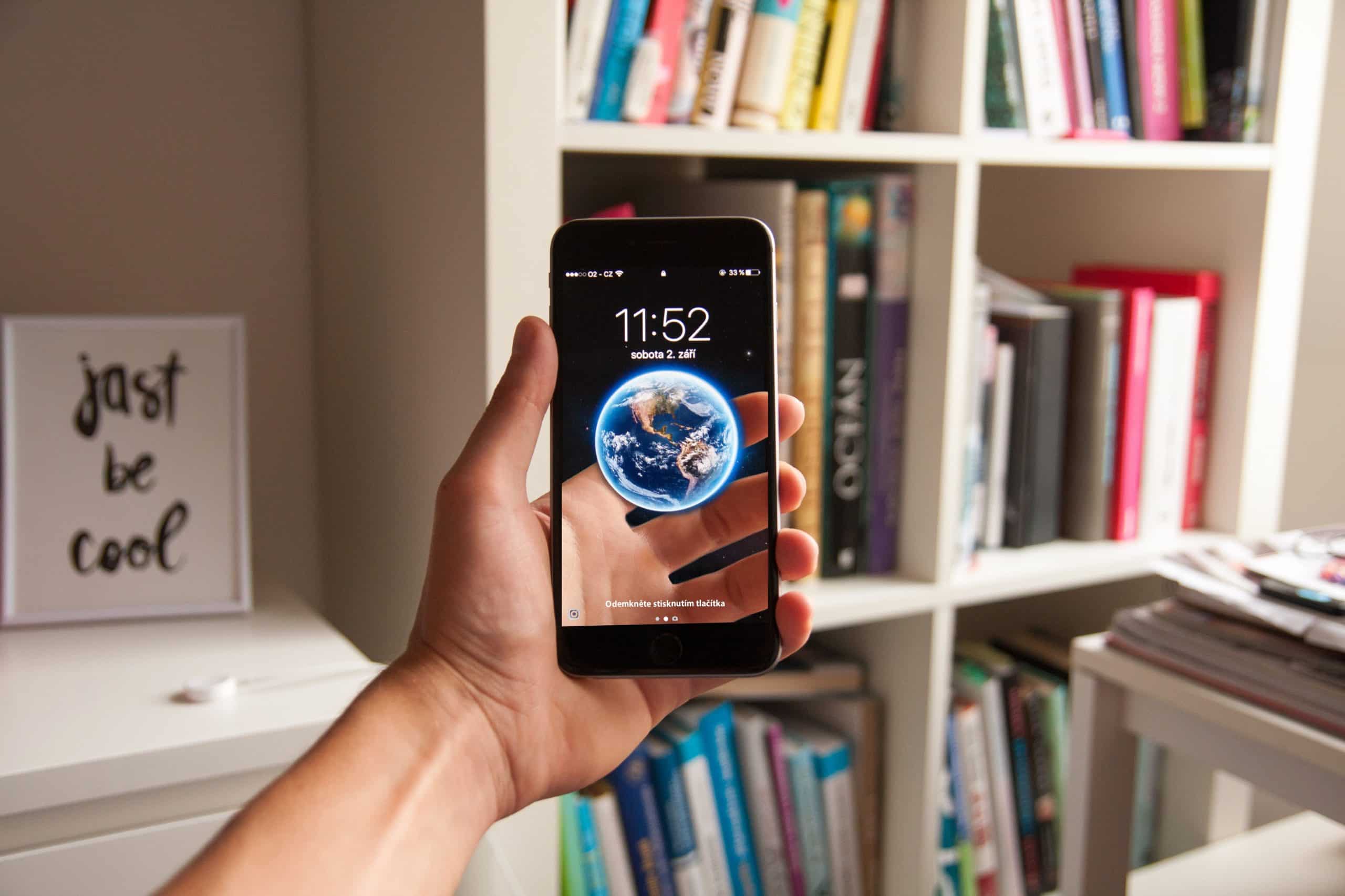 A person holding an iPhone 9 displaying the time 11:52 and an image of Earth as the wallpaper. The background shows bookshelves filled with various books and a framed sign that reads "just be cool.