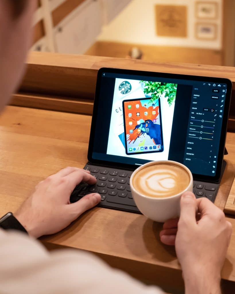 A person holding a cup of coffee while working on an iPad Pro with a keyboard. The tablet screen displays a graphic design editing app. The wooden table and cozy indoor setting suggest a workspace or café environment, perfect for utilizing the 5G-enabled device's capabilities.