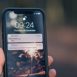 A close-up of a hand holding an iPhone displaying the lock screen. The time is 09:24 on Thursday, December 28. Notifications shown include a WhatsApp message from "Oscar Sutton", a Twitter notification about "Luke Chesser" liking a photo on Unsplash, and an update from Facebook.