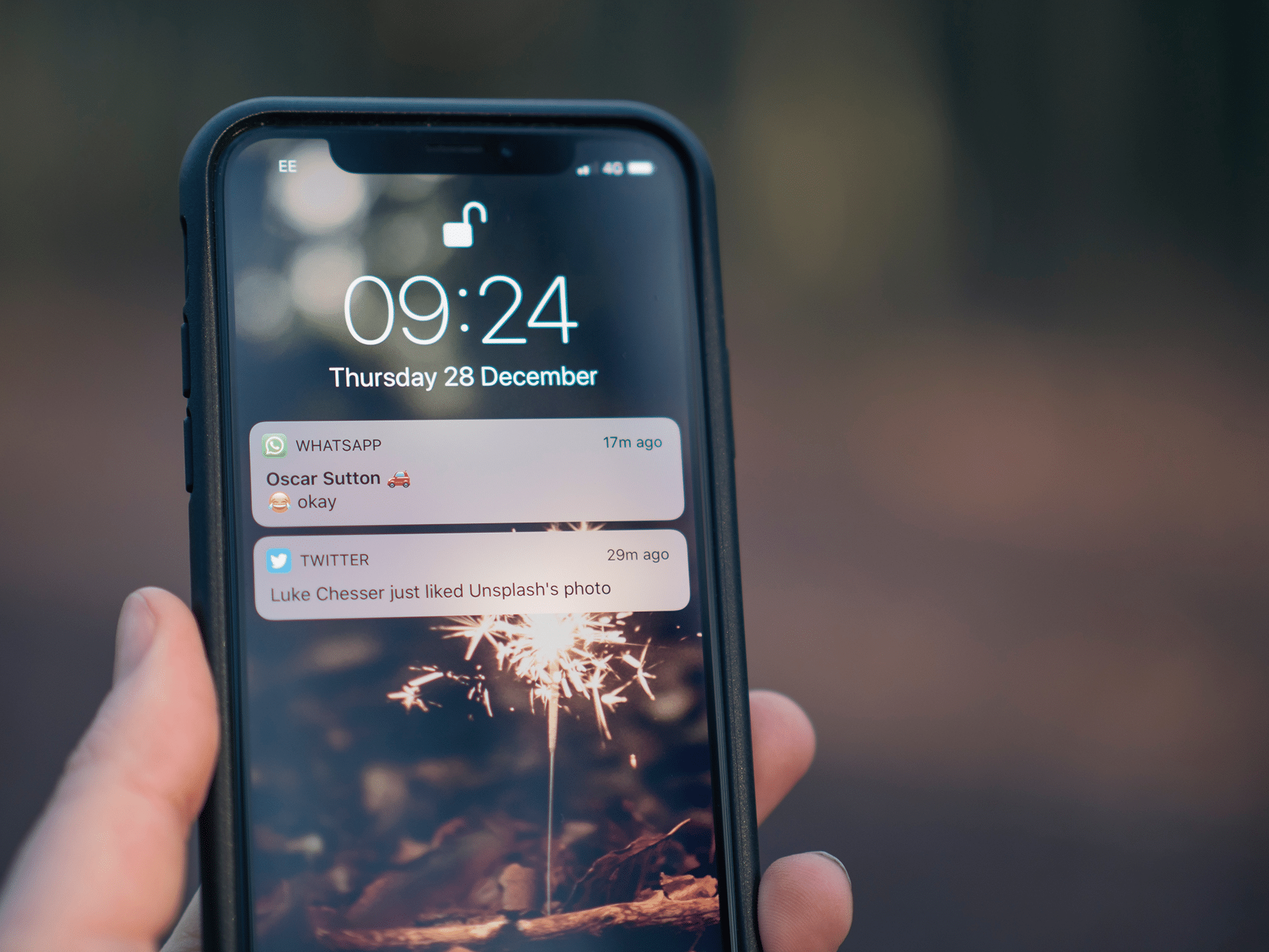 A close-up of a hand holding an iPhone displaying the lock screen. The time is 09:24 on Thursday, December 28. Notifications shown include a WhatsApp message from "Oscar Sutton", a Twitter notification about "Luke Chesser" liking a photo on Unsplash, and an update from Facebook.