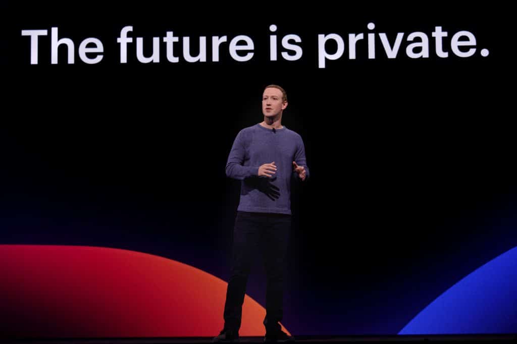 A person stands on stage in front of a large screen displaying the text "The future is private." They are wearing a long-sleeve shirt and dark pants, with abstract red and blue shapes behind them. The event highlights Facebook's spring announcement of its redesigned desktop site focused on enhanced privacy.