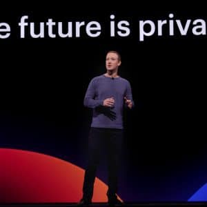 A person stands on stage in front of a large screen displaying the text "The future is private." They are wearing a long-sleeve shirt and dark pants, with abstract red and blue shapes behind them. The event highlights Facebook's spring announcement of its redesigned desktop site focused on enhanced privacy.