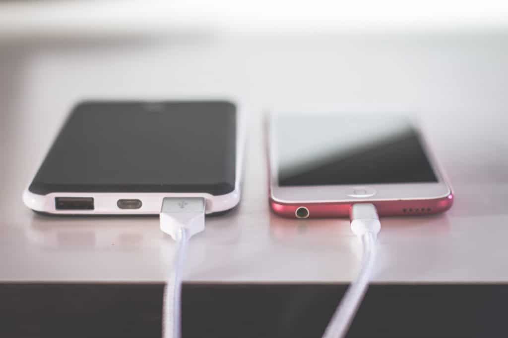 Two smartphones are placed side by side on a white surface, both connected to white charging cables. The phone on the left, possibly an Apple model, has a black and white case, while the one on the right has a red case. Both devices appear to use an EU standardized charging port.
