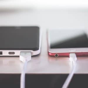 Two smartphones are placed side by side on a white surface, both connected to white charging cables. The phone on the left, possibly an Apple model, has a black and white case, while the one on the right has a red case. Both devices appear to use an EU standardized charging port.