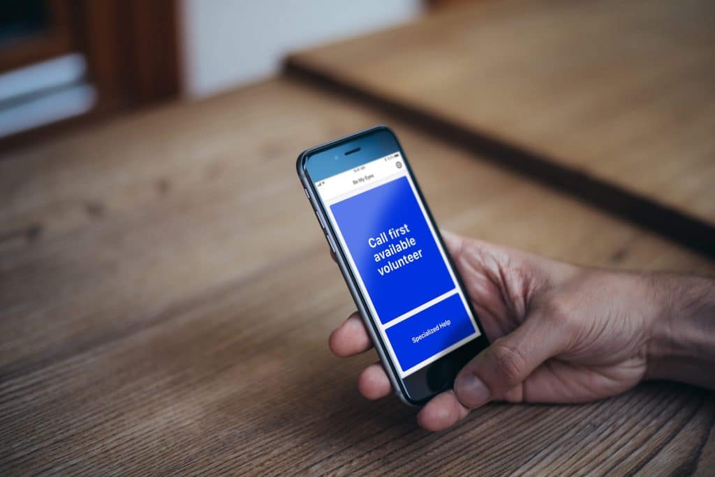 A hand holding a smartphone displaying a blue screen with white text that reads "Call first available volunteer." The phone is on a wooden table, and the background is slightly blurred, showcasing the Be My Eyes app for sighted volunteers aiding blind people.