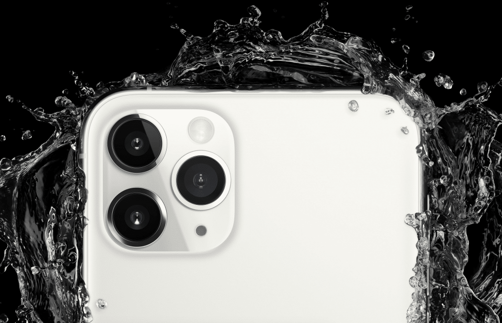 Close-up of the top corner of a white smartphone with three camera lenses. Water splashes around the phone, highlighting its water resistance feature against a black background. Despite challenges like Coronavirus affecting iPhone shipments, this model stands out with its durability.