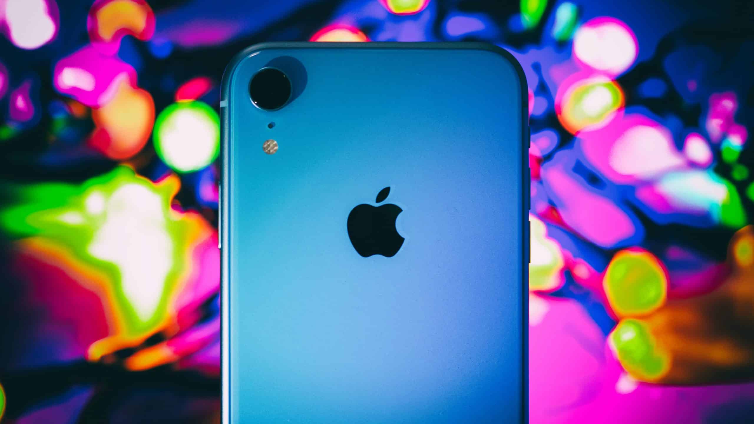 A close-up of a blue iPhone XR showing the Apple logo and camera on the back. The background is blurred with vibrant, colorful lights in shades of pink, green, yellow, and purple, creating a bokeh effect. This 2019 model remains a popular smartphone choice for many.