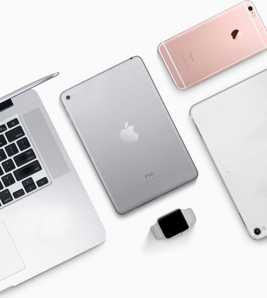 A flat lay image of several Apple devices running iOS and macOS displayed on a white surface, including a MacBook, an iPad, an Apple Watch, a rose gold iPhone, and a silver iPhone. These devices exemplify the seamless integration of Universal Purchases across the Apple ecosystem.