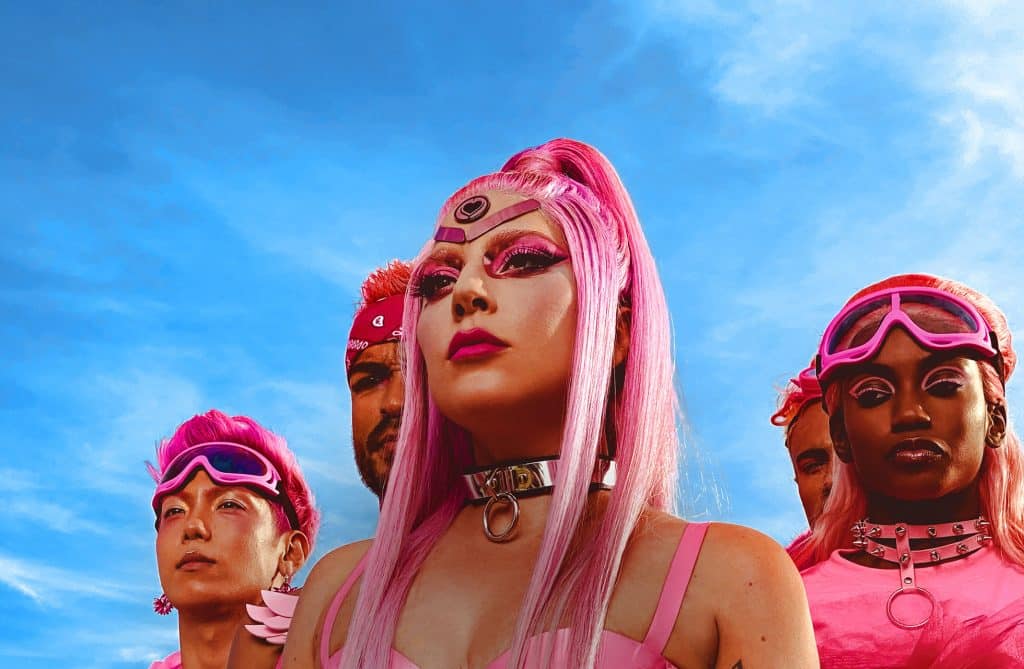 A group of individuals with vibrant pink hair and accessories stand confidently under a blue sky. The central figure, reminiscent of Lady Gaga, wears a headpiece and bold pink makeup, while the others, all set for a music video shoot on an iPhone 11, are adorned with goggles and bandanas, looking determined.