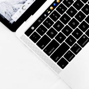 A close-up of a 2021 ARM MacBook keyboard, featuring a touch bar displaying various emoji icons such as smiling faces, thumbs up, and hearts. The background is minimalistic with a white table surface. The upper portion shows the edge of the Apple laptop screen.