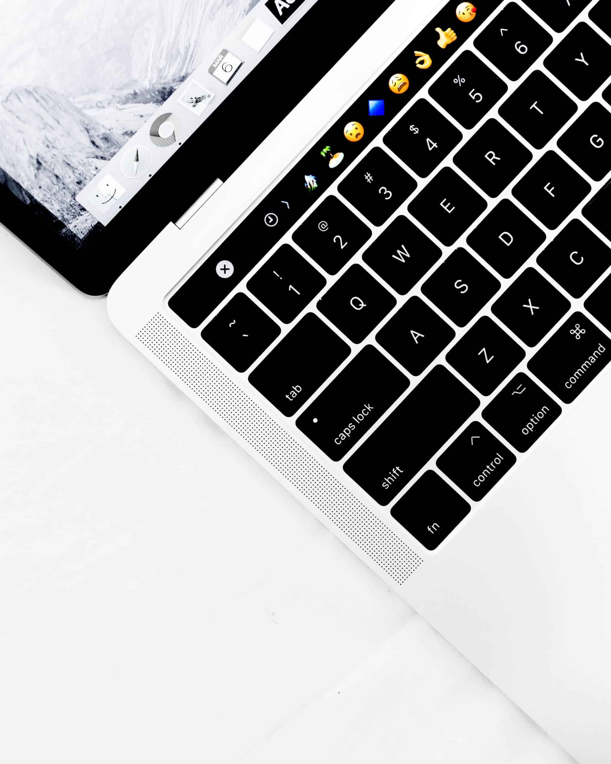 A close-up of a 2021 ARM MacBook keyboard, featuring a touch bar displaying various emoji icons such as smiling faces, thumbs up, and hearts. The background is minimalistic with a white table surface. The upper portion shows the edge of the Apple laptop screen.