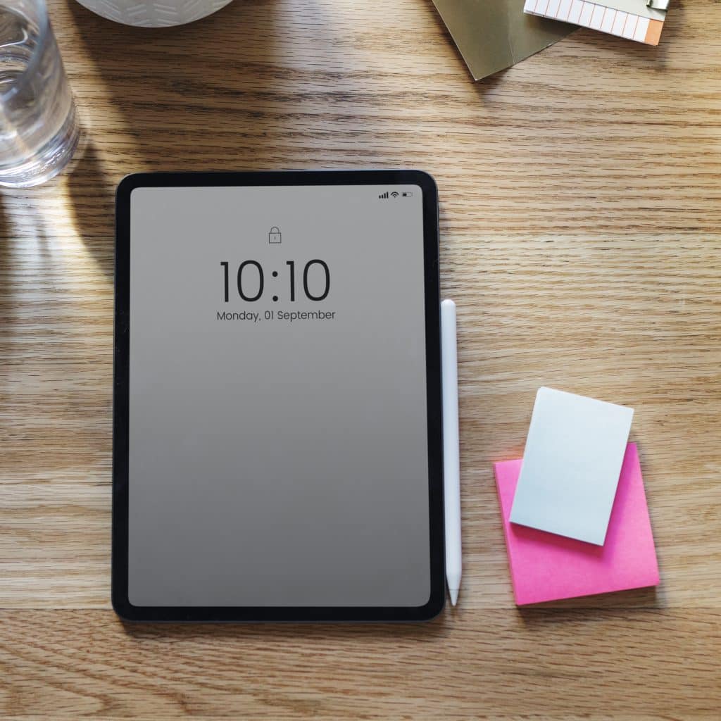 An iPad displaying 10:10 AM on its lock screen lies on a wooden desk, connected to an Apple Smart Keyboard. Nearby are a white stylus and a small stack of sticky notes in pink and blue. A glass of water and some stationery items complete the organized workspace.